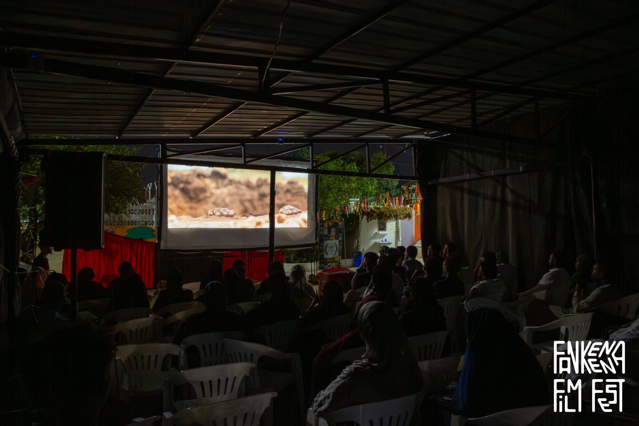The audience watches a film on the big screen under the courtyard roof at night